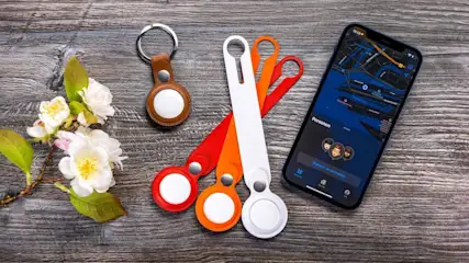 Auf einem Holztisch liegen AirTags, Schlüsselanhänger, Blütenzweig und ein Smartphone mit Karten-App.