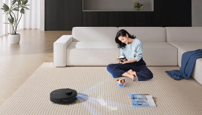 Eine Frau sitzt auf einem Teppich mit einem Roboterstaubsauger, einer Schale Obst und einer Zeitschrift.