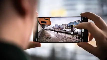 Eine Person hält ein Smartphone, um eine Uferpromenade mit Gebäuden und Wasser im Hintergrund zu fotografieren.