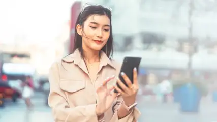 Eine Person mit Hemd hält ein schwarzes Handy im Freien. Im Hintergrund sind verschwommene Gebäude und Passanten.