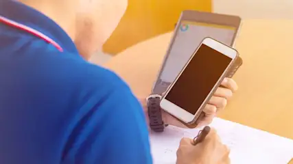 Eine Person in blauem Hemd hält ein Smartphone und einen Stift über einem Blatt Papier. Ein zweites Smartphone steht im Hintergrund.