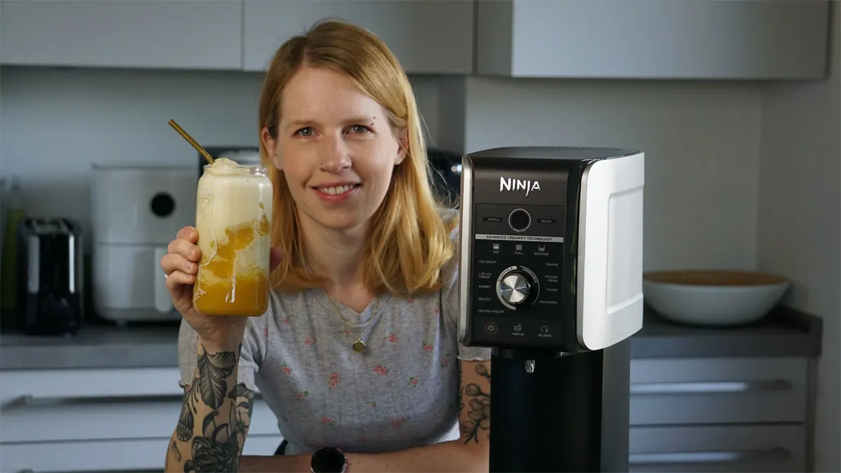 Franzi steht neben der Ninja Creami Deluxe und hält einen Mango-Matcha-Frappé in der Hand.