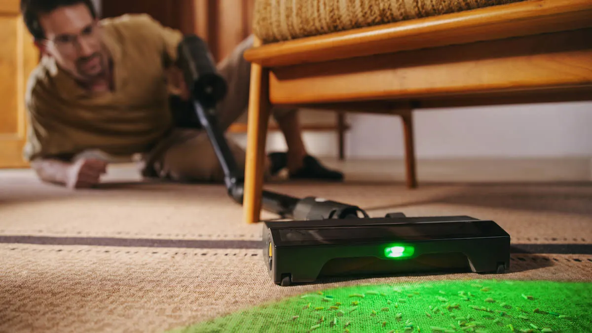 Ein Mann saugt mit dem Roborock H60 Ultra unter einem Sofa, der Staubsauger wirft grünes Licht auf den Teppichboden.