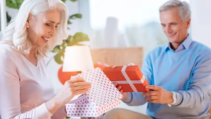 Eine Frau öffnet ein Geschenk mit Herzmuster, während ein Mann ein rotes Geschenk mit weißem Band hält.