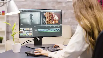 Frau an Schreibtisch vor einem Monitor, an dem ein Videoschnittprogramm mit zwei Ausschnitten, den Video- und Audiospuren sowie eine Bibliothek darauf zu sehen sind.