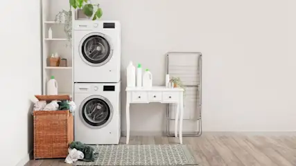 Ein Waschraum mit weißer Waschmaschine und Trockner, einem gefüllten Wäschekorb, einem Regal und einem Tisch mit Reinigungsmitteln.