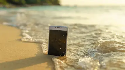 Ein Smartphone mit schwarzem Bildschirm steht im Sand, umspült von hellen Wellen am Ufer.