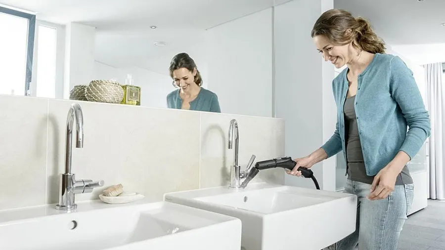 Eine Frau reinigt ein weißes Waschbecken mit einem Dampfreiniger in einem hellen Badezimmer mit Spiegel.