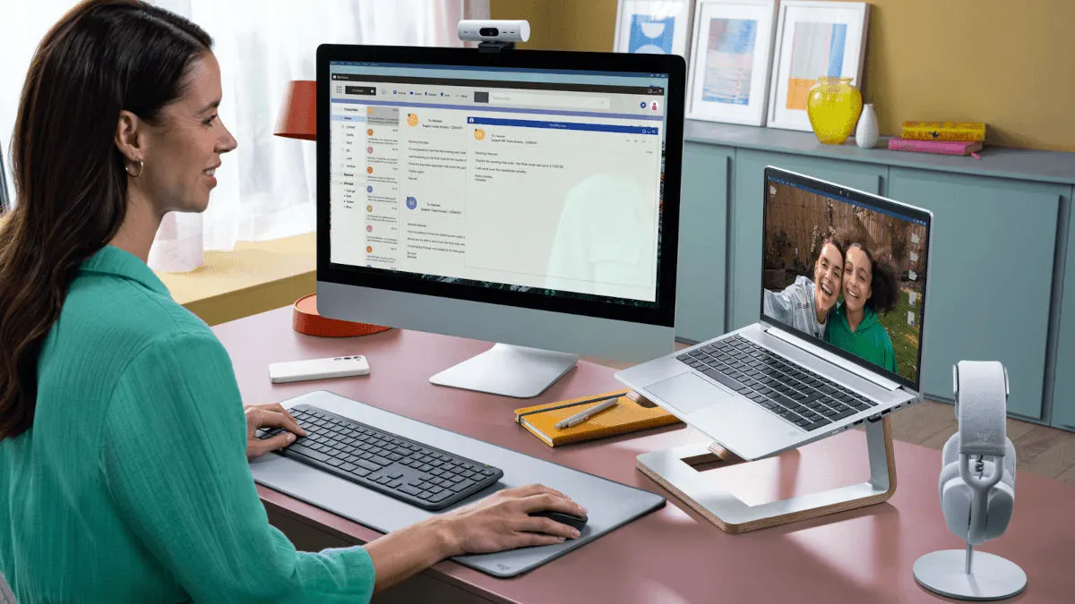 Eine Frau arbeitet an einem Schreibtisch mit Computern, Tastatur, Maus und Kopfhörern. Auf einem Laptop ist ein Videoanruf zu sehen.