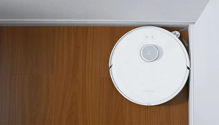 Ein runder, weißer Staubsaugerroboter steht auf einem braunen Holzboden neben einer weißen Wand und einer Tür.