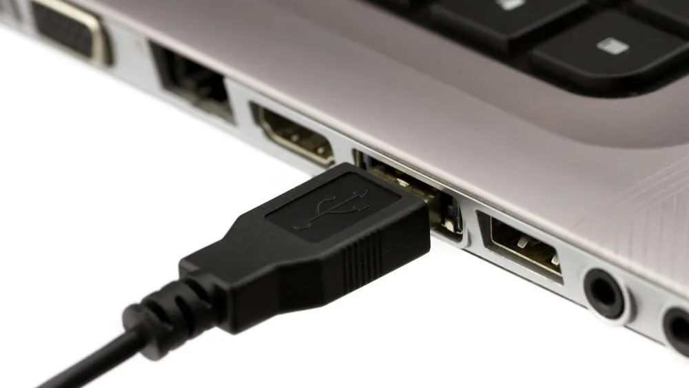 Ein schwarzer USB-Stecker ist an der Seite eines silbernen Laptops angeschlossen, neben anderen Anschlüssen und schwarzen Tasten.