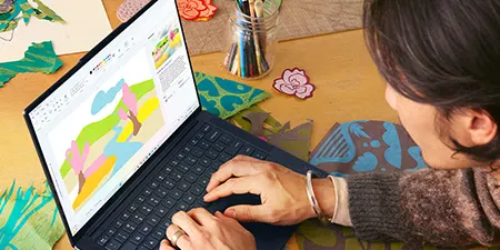 Eine Person tippt auf einem Laptop, der eine farbige Grafik anzeigt. Auf dem Tisch liegen Bastelmaterialien und Werkzeuge.