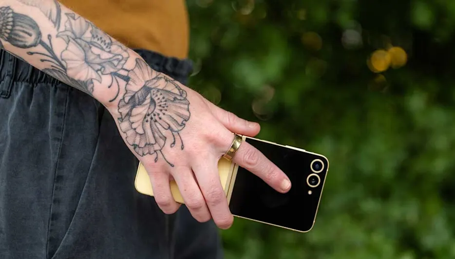 Eine tätowierte Hand hält ein schwarzes und goldenes Smartphone vor grünem Hintergrund. Die Person trägt einen goldenen Ring.