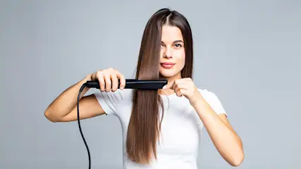 Eine Frau in Weiß glättet ihr langes, braunes Haar mit einem schwarzen Glätteisen vor einem grauen Hintergrund.