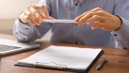 Eine Person fotografiert mit einem Smartphone ein Diagramm auf einem Klemmbrett. Laptop und Stift liegen auf einem Holztisch.