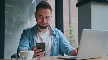 Ein Mann sitzt hält ein Smartphone in der Hand und sitzt vor einem Notebook