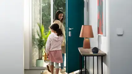 Mutter und Kind betreten ein Haus durch eine offene blaue Tür. Eine Lampe und ein Lautsprecher stehen auf einem Tisch.