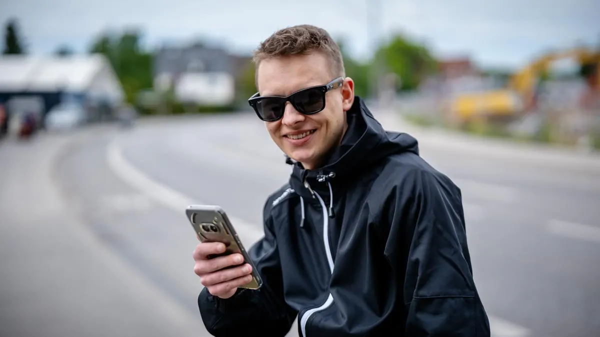 Ein Mann mit Sonnenbrille und schwarzer Jacke hält ein Smartphone auf einer grauen Straße. Im Hintergrund sind Gebäude und Bäume.
