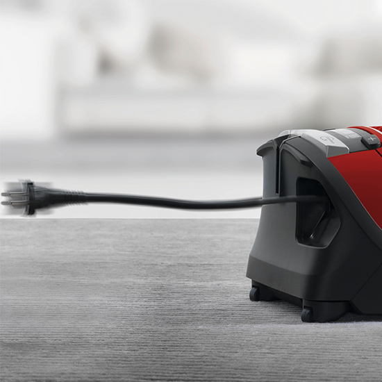 Ein Staubsauger in Rot und Grau mit einem schwarzen Kabel auf einem grauen Untergrund vor einem verschwommenen Hintergrund.