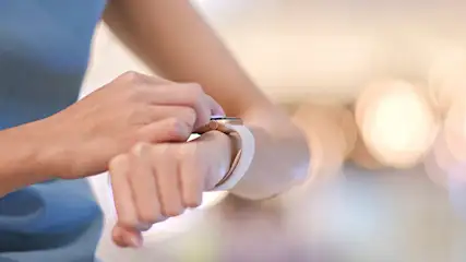 Eine Person mit einem blauen Oberteil bedient eine Smartwatch mit einem hellen Armband. Der Hintergrund ist verschwommen.