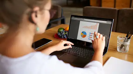 Eine Person arbeitet an einem Laptop mit Diagrammen auf einem Holztisch. Daneben liegen Früchte, ein Smartphone und Büromaterial.