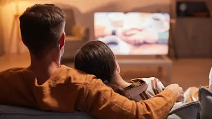 Ein Paar sitzt auf einem grauen Sofa und schaut auf einen Fernseher in einem Wohnzimmer mit warmem Licht.