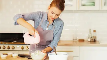 Eine Frau mit Schürze mixt Teig in einer Glaschüssel mit einem rosa Handmixer in einer hellen Küche. Im Hintergrund stehen Schränke.