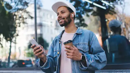 Ein Mann mit Kappe hält ein Smartphone und einen Kaffeebecher im Freien. Im Hintergrund sind Bäume und Gebäude.