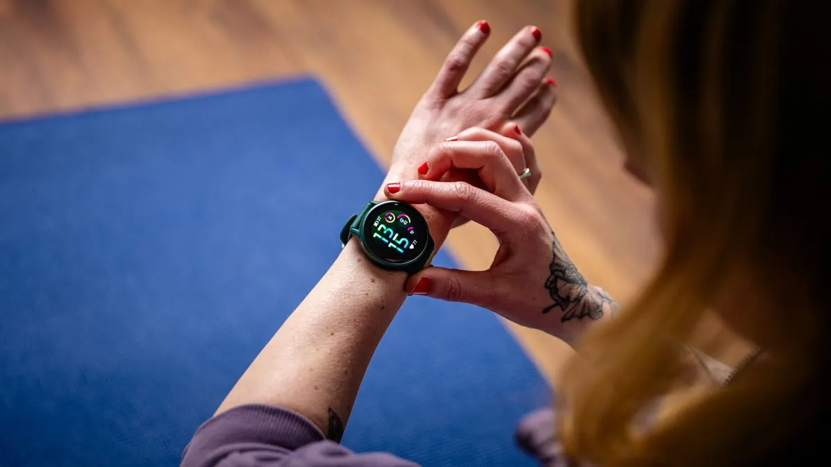 Eine Frau mit rotem Nagellack betrachtet eine grüne Smartwatch am Handgelenk vor einer blauen Matte.