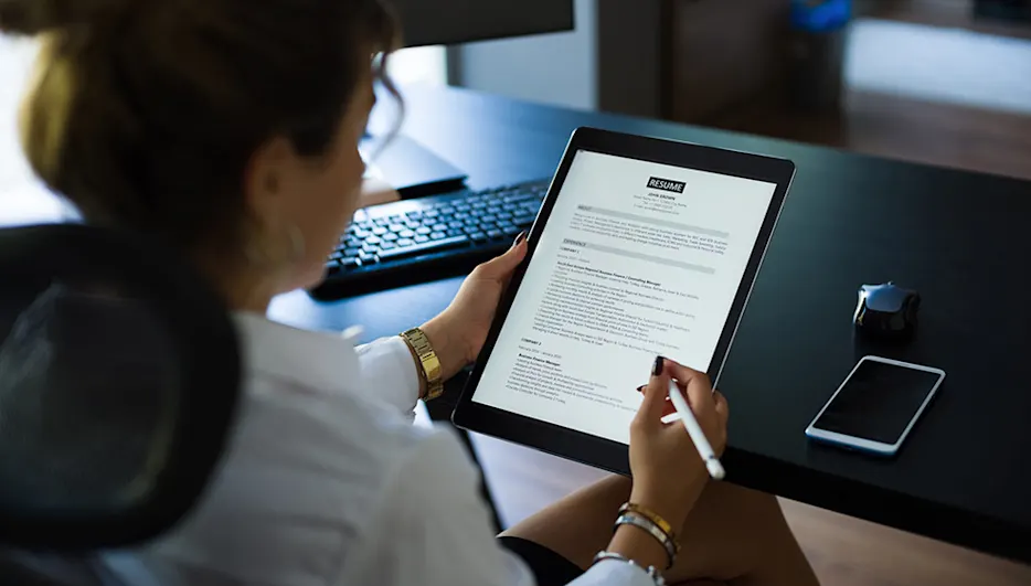 Frau betrachtet Lebenslauf auf Tablet mit Stift. Tastatur, Handy und Büromaterial auf schwarzem Tisch im Hintergrund.