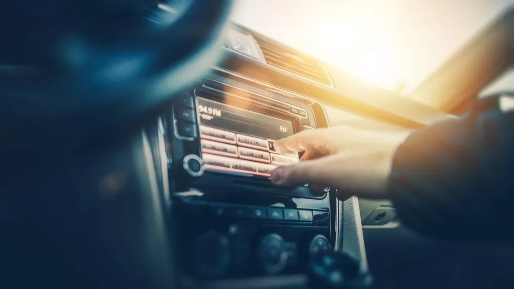 Eine Hand bedient das Autoradio in einem Auto. Das Radio hat Knöpfe und einen Bildschirm mit der Frequenzanzeige.