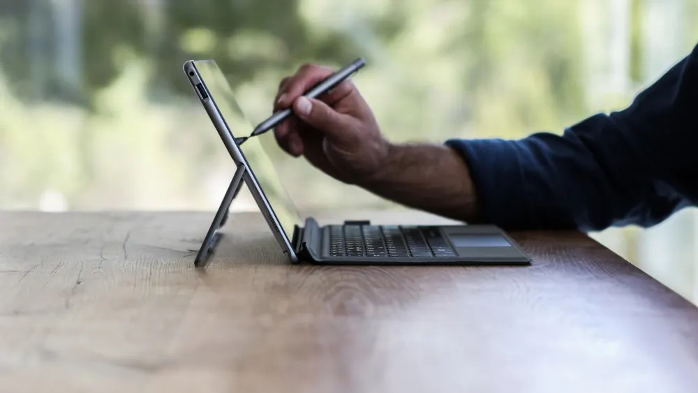 Ein Tablet steht auf einem Holztisch, ein Stift wird von einer Hand auf dem Bildschirm gehalten, mit Tastatur und grünem Hintergrund.