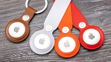 Nahaufnahme von vier Apple AirTags in braunen, grauen, orangen und roten Halterungen auf einem grauen Untergrund.