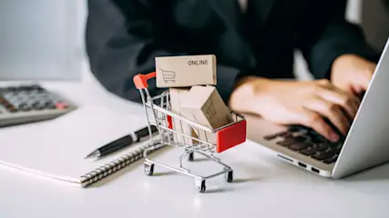 Ein Mini-Einkaufswagen mit Kisten, die "Online" zeigen, steht neben einem Laptop, Notizbuch, Stift und Rechner auf einem Tisch.