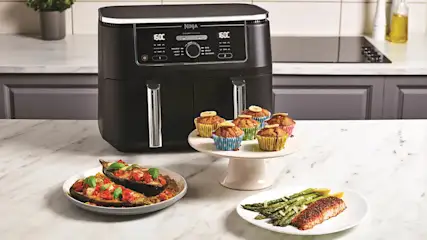 Die Heißluftfritteuse Ninja AF400EU in einer hellen Küche. Vor der Fritteuse stehen verschiedene Speisen.