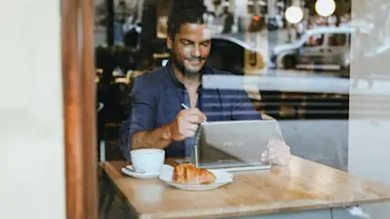 Mann an Tisch mit Tablet, Gebäck und Kaffee. Im Hintergrund Stadtbild durch Fensterglas sichtbar.