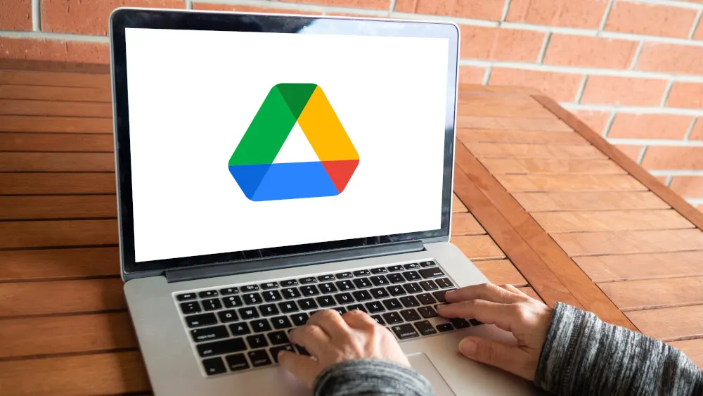 Person tippt auf einem Laptop mit Google Drive-Logo auf dem Bildschirm, der auf einem Holztisch vor einer Backsteinmauer steht.