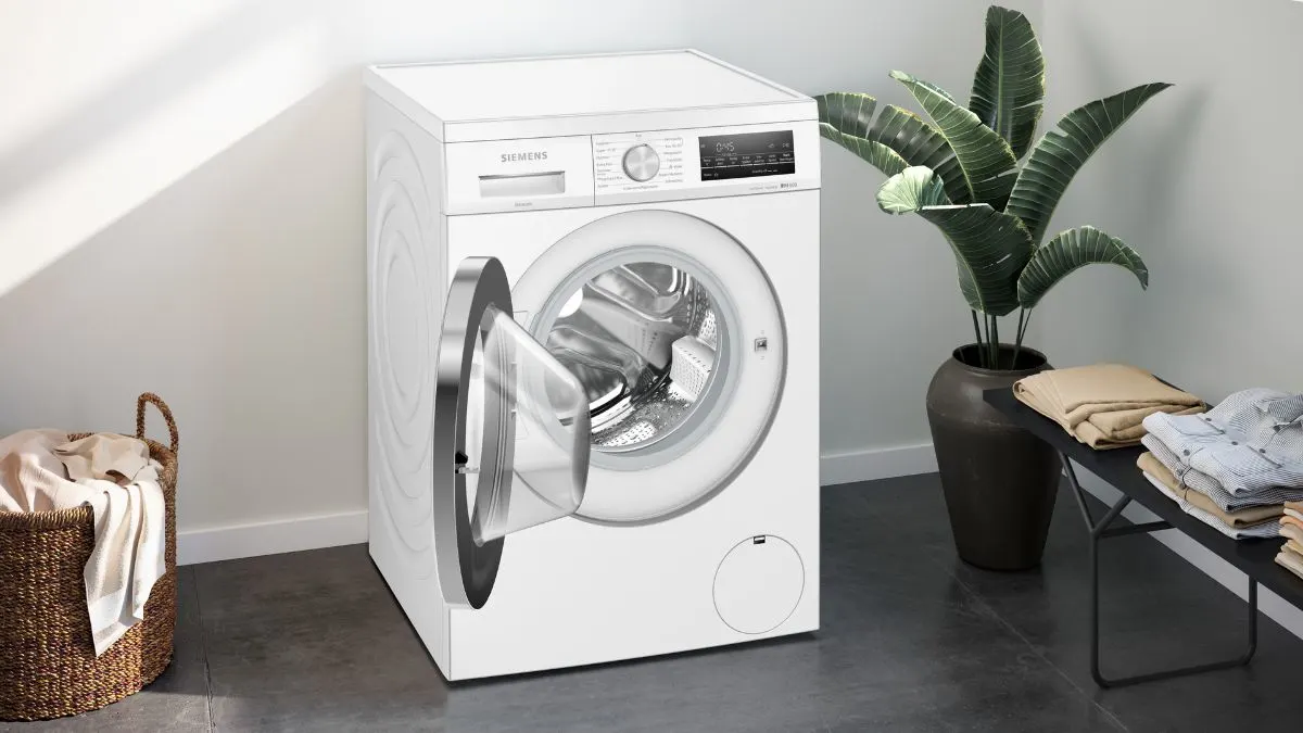 Eine weiße Siemens Waschmaschine mit offener Tür steht neben einem Korb mit Wäsche. Im Hintergrund eine Pflanze und ein Tisch mit Kleidung.