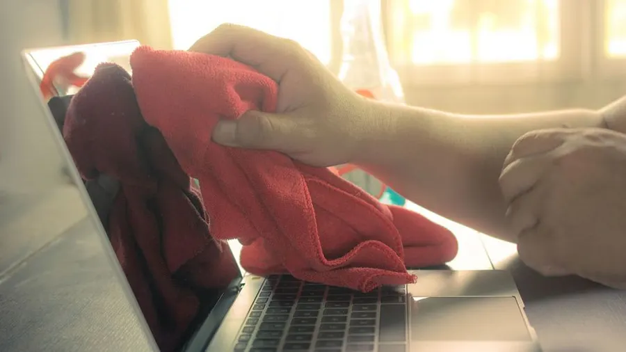 Eine Hand wischt mit einem roten Tuch über die Tastatur eines Laptops. Im Hintergrund ist ein Fenster sichtbar.