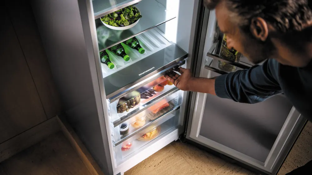 Ein Mann öffnet einen Kühlschrank mit Lebensmitteln auf mehreren Regalen, darunter Salat, grüne Flaschen und verpacktes Fleisch.