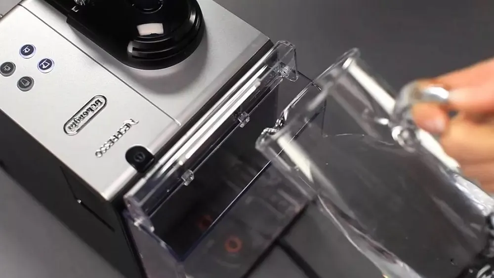 Silberne Nespresso-Kaffeemaschine mit Tasten, Wassertank und einer Hand, die einen Glasbecher hält.