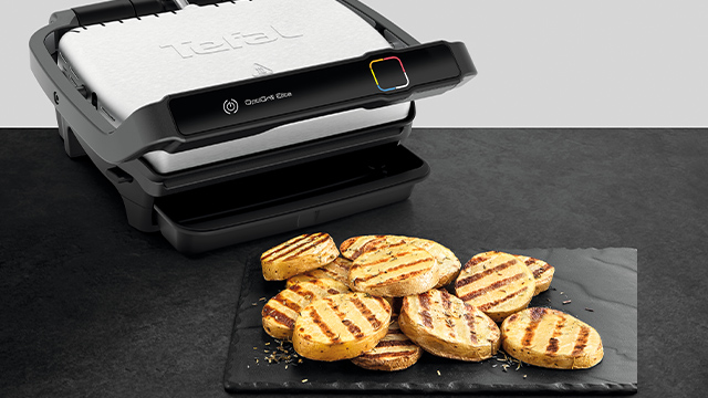 Ein Grill mit gegrillten Kartoffelscheiben auf einem schwarzen Teller. Der Hintergrund ist grau und weiß.