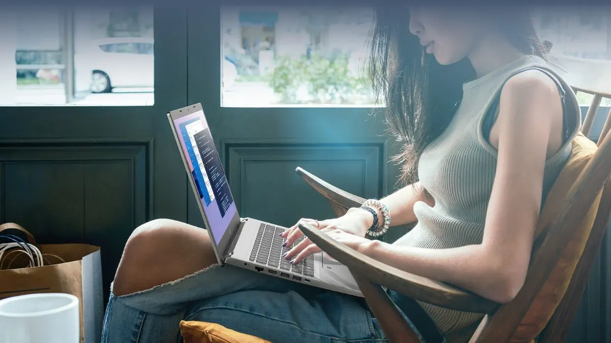 Frau sitzt in einem Holzstuhl und tippt auf einem Laptop. Ein Fenster und eine Papiertüte sind im Hintergrund sichtbar.