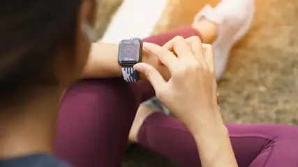 Frau in Sportkleidung tippt auf eine Smartwatch mit Herzfrequenzanzeige. Im Hintergrund sind ein Schuh und eine Bodenfläche zu sehen.