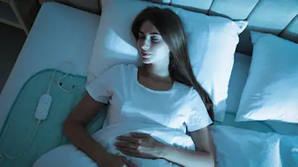 Eine Frau schläft in einem weißen T-Shirt in einem Bett mit weißen Kissen und einer blauen Heizdecke.