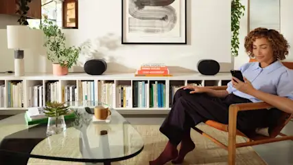 Frau sitzt in einem Holzstuhl und benutzt ein schwarzes Handy. Im Hintergrund sind Bücherregale mit Büchern und zwei Lautsprechern.