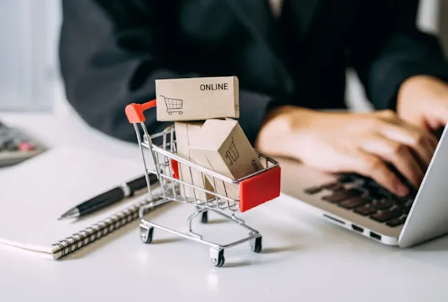 Ein Mini-Einkaufswagen mit Kartons, die "Online" und ein Warenkorb-Symbol tragen, steht neben einem Laptop, Notizbuch und Stift.