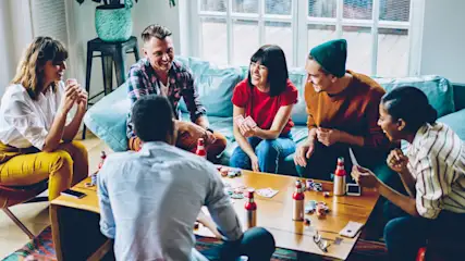 Eine Gruppe von sechs Leuten sitzt und spielt Karten an einem Tisch in einem Wohnzimmer. Es stehen Getränke und Chips auf dem Tisch.