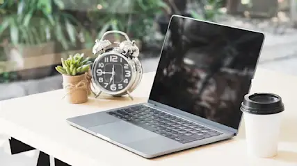 Ein Laptop, ein Wecker, eine Topfpflanze und ein Kaffeebecher stehen auf einem Tisch vor einem Fenster.