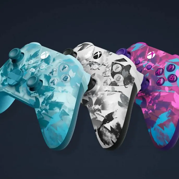 XBOX Series X 1TB Drei Xbox‑Controller im Camouflage‑Design (türkis, schwarz‑weiß, pink/blau) nebeneinander, kabellose Controller mit Analogsticks, Steuerkreuz und A/B/X/Y‑Tasten.